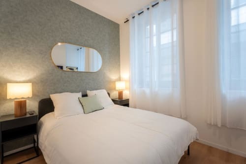 Modern bedroom in Bordeaux apartment with white bedding, wooden nightstands, oval mirror, and sheer white curtains