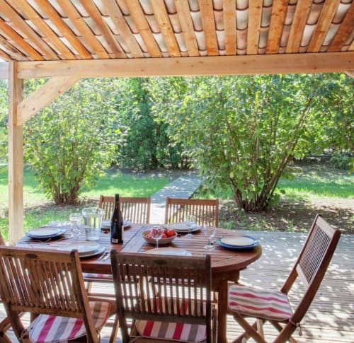 Zona pranzo all'aperto presso Le Maquis Plage con pergolato in legno, sedie a righe e vista su rigogliosi giardini a Poggio-Mezzana.