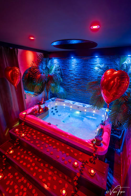 Spa privatif luxueux avec bain à remous aux lumières néon rose et bleu, ballons cœur et plantes tropicales à L'Insolite Spa Privatif