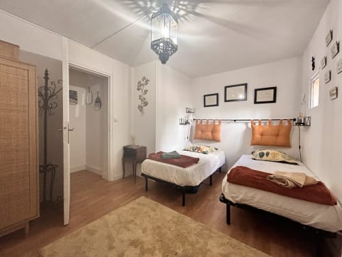 Chambre lumineuse avec deux lits, coussins orange, luminaire suspendu, parquet et murs blancs dans un appartement d'Arles