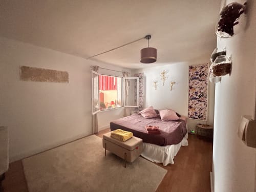 Chambre douillette dans un appartement d'Arles avec lit violet, fenêtre aux volets rouges, luminaire suspendu et décoration florale murale