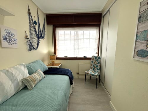 Dormitorio luminoso en apartamento de Baiona con vistas al mar, ropa de cama azul claro, decoración costera y gran ventana con vistas a la bahía