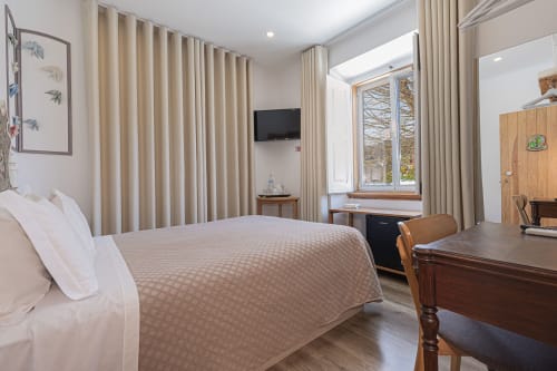 Spacious hotel room at Ninho Saloio with queen bed, beige curtains, wooden desk, TV, large windows overlooking garden,