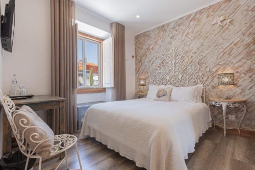 Quarto padrão elegante no hotel Ninho Saloio com parede de destaque estampada, roupa de cama branca, mesa de madeira e vista para a cidade.