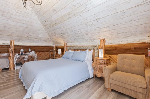 Rustic wooden bedroom at Ninho Saloio with blue bedding, log posts, tan armchair, and white vaulted ceiling