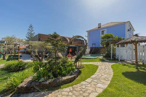 Pátio do hotel Ninho Saloio em Venda do Pinheiro com casa azul, caminhos de pedra, jardins frondosos e estruturas de madeira