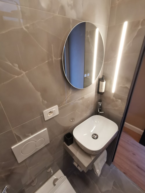 Moderno bagno dell'Hotel Fiori Roma con specchio LED ovale, lavabo bianco e piastrelle grigie