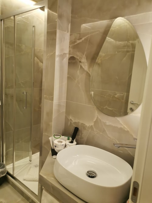 Bagno moderno dell'Hotel Fiori Roma con lavabo ovale bianco, specchio tondo, box doccia in cristallo e piastrelle con motivo marmoreo