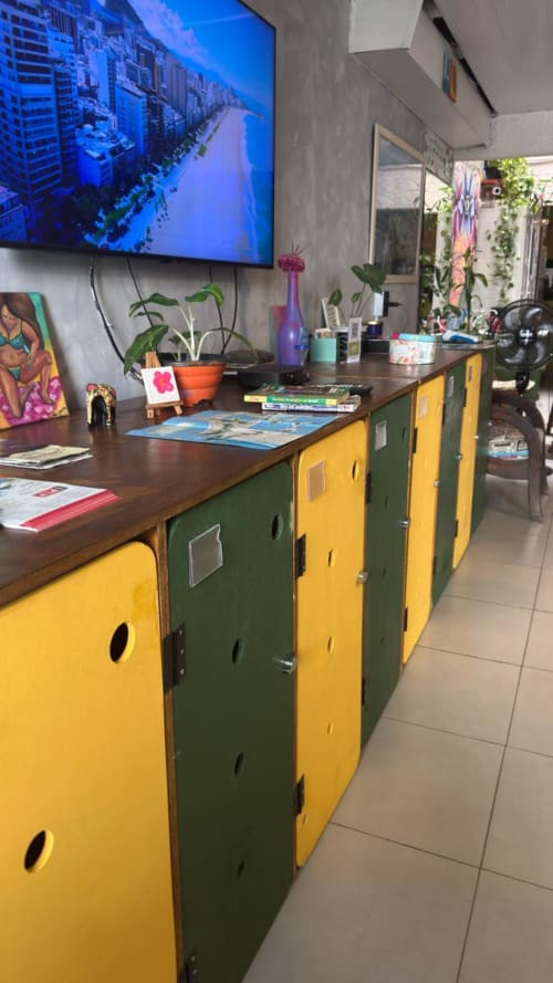 Área comum colorida de albergue com armazenamento amarelo e verde, mesa de madeira, plantas e TV exibindo paisagem do Rio
