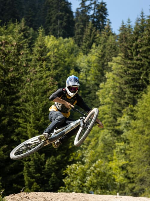 Vététiste en vol au-dessus d'un saut en terre, forêt de pins en arrière-plan à Montgenèvre