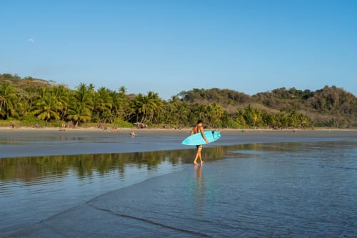 Surfer with blue board wading in calm ocean waters at Paquera beach, Costa Rica, near Organica Yoga Shelter