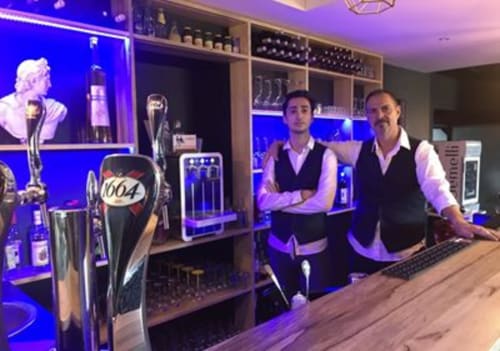 Twee personeelsleden van bar La Maison D'ambrine in Chamaret, Frankrijk, met blauwe neonverlichting en biertap-displays.