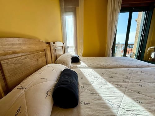 Dormitorio soleado en villa Vila Arume, Baiona, con camas gemelas, cabeceros de madera y vistas al océano desde el balcón
