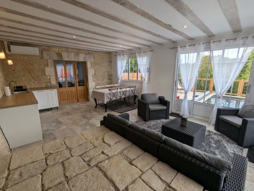 Petit Salon du gîte La Grange des Colporteurs avec murs en pierre, poutres en bois, mobilier en osier, coin cuisine, et vue sur piscine et jardin. Gîte de charme en Charente, idéal pour un séjour nature et détente.