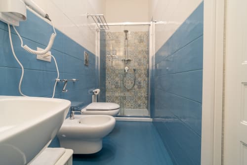 Bagno moderno con piastrelle blu alle pareti, lavandino bianco, toilette, bidet e box doccia in vetro presso Le Boutique del Centro