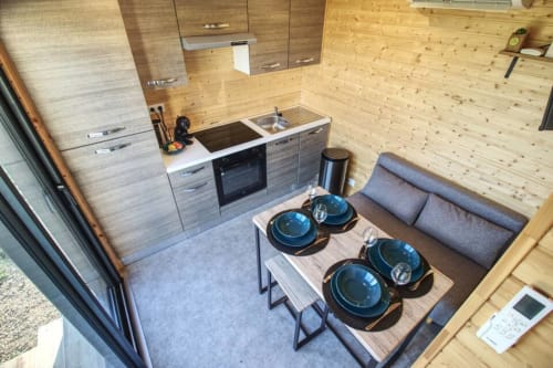Compact kitchen-living area with wooden walls, dining table set with blue dishes, gray sofa, and modern appliances at Ilot