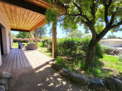 Covered wooden deck terrace at Villas Lantana in Lecci with outdoor seating, mature tree, and lush garden views