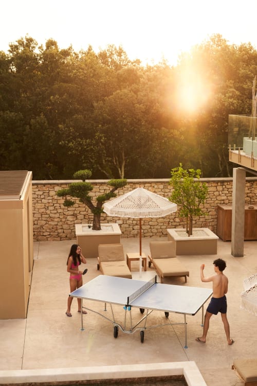 Dos niños jugando al tenis de mesa en la soleada terraza de la villa La Maison D'ambrine en Chamaret, con tumbonas y árboles