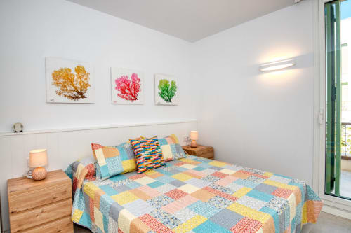 Dormitorio colorido en Finques Garcia Enrich de Palafrugell con colcha patchwork, mesillas de madera y arte estacional con árboles.