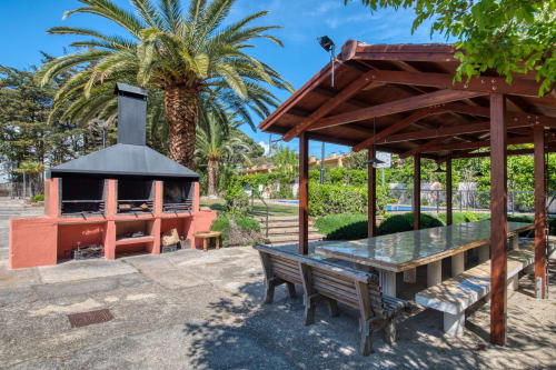 Outdoor patio area with red brick BBQ grill, wooden pergola, dining table, and palm trees at Finques Garcia Enrich in