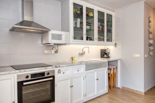 Modern white kitchen with marble countertop, built-in oven, range hood, and glass-fronted cabinets at Finques Garcia Enrich