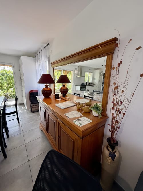 Angolo credenza con mobile in legno e grande specchio nell’area soggiorno del gîte, decorato con lampada rossa e vaso ornamentale.