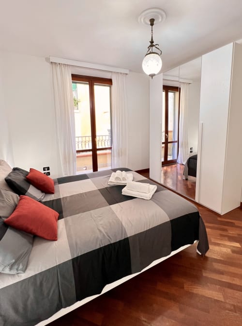 Ampia camera da letto di LA Home Viareggio con biancheria a righe, cuscini rossi, accesso al balcone, armadio con specchio e elegante lampada a sospensione