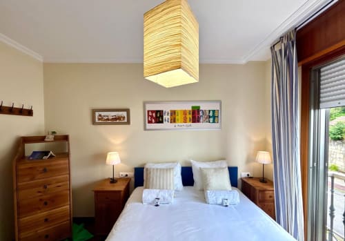 Dormitorio luminoso en apartamento Chuliña, Baiona con cama doble, muebles de madera, lámpara colgante rayada y acceso a balcón
