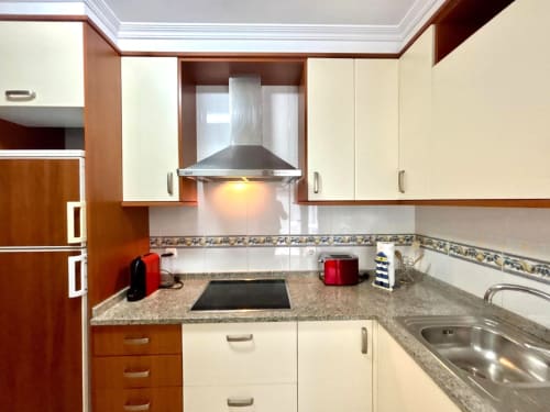 Cocina moderna con muebles crema, encimeras de granito, placa vitrocerámica de acero inoxidable y campana extractora en apartamento de Chuliña, Baiona