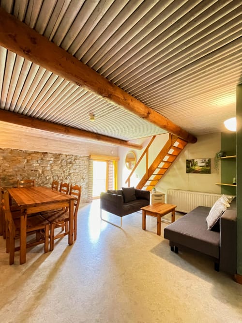 Intérieur spacieux de La Ferme De Werpin avec poutres en bois, table à manger, canapé noir et escalier en bois