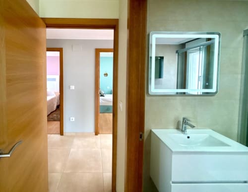 Baño moderno con lavabo blanco, espejo iluminado y puertas abiertas hacia el dormitorio en la casa cerca de Playa América