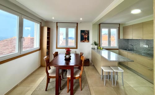Cocina y comedor modernos en la casita Laranxeira, Nigrán, con vistas al mar a través de grandes ventanales, mesa de madera y luz natural