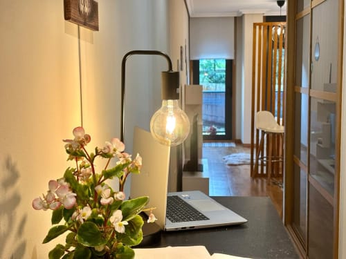 Acogedor espacio de trabajo en apartamento de Baiona con portátil, plantas en maceta y lámpara colgante con vista al pasillo hacia la piscina
