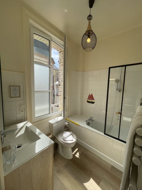 Modern bathroom at Hotel Le Trouville with bathtub, shower, sink, and city views of Trouville sur Mer through window