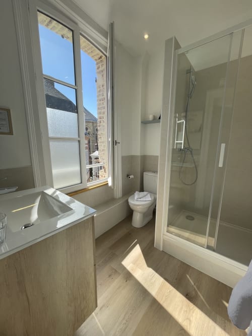 Modern bathroom at Hotel Le Trouville with sink, toilet, glass shower enclosure, and city view window overlooking Trouville