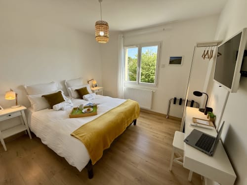 Camera Gex del gîte, letto queen size con biancheria bianca e coperta ocra, scrivania con laptop e finestra vista giardino.