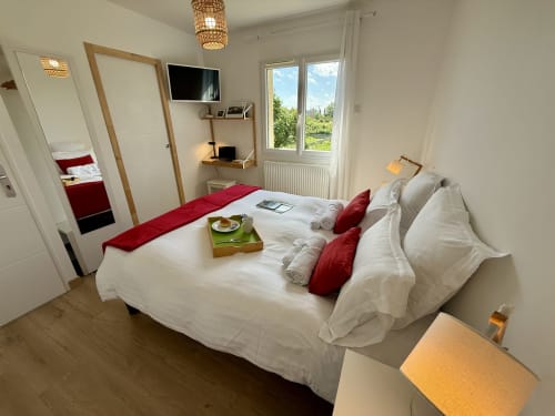 Bresse room at the guesthouse featuring a double bed with white linens and red cushions, wall-mounted wooden desk, TV, and window overlooking the garden.