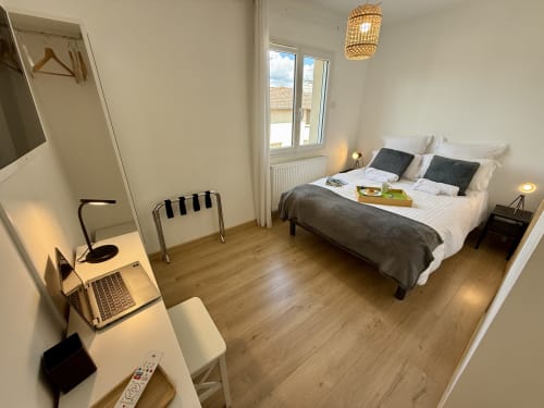 Camera Bugey del gîte con letto matrimoniale con copriletto grigio, cuscini blu, scrivania a muro bianca, TV a schermo piatto e armadio aperto.
