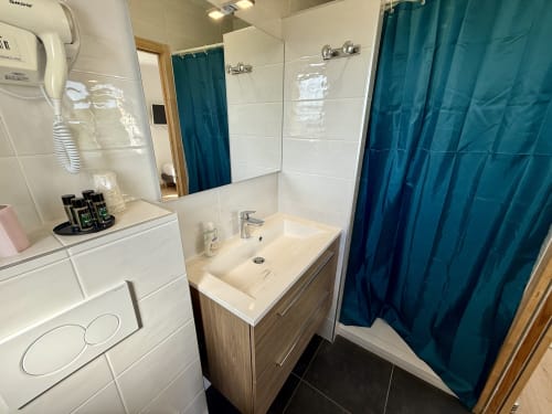 En-suite bathroom of the Dombes room, featuring a blue shower curtain, wooden vanity unit and mirror reflecting the bedroom.