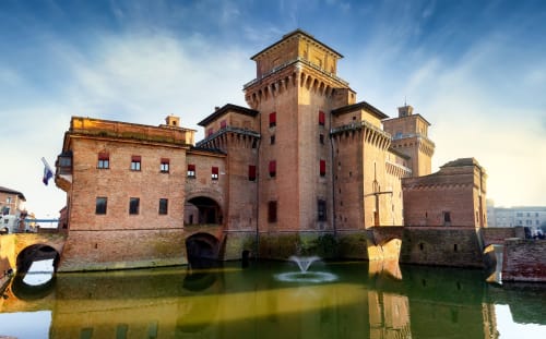 Medieval brick fortress on moat in Ferrara, Italy with blue sky reflecting in water
