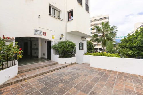 Marbella apartment entrance courtyard with terracotta tile patio, white buildings, tropical plants, and sea view in Puerto
