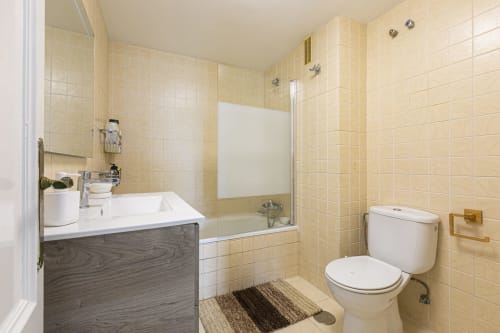 Modern bathroom in Marbella apartment with beige tile walls, vanity sink, bathtub with shower screen, and toilet