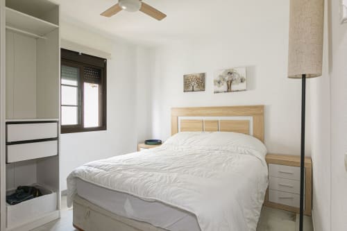 Bright bedroom in Marbella apartment with wooden bed frame, white bedding, ceiling fan, and sea-view wall art