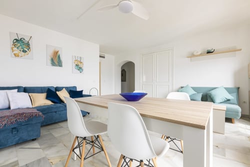 Modern open-plan living area in Puerto Banús apartment with blue sofas, dining table, white chairs, and botanical wall art.