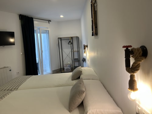 Moderno dormitorio de apartamento en Mérida con cama blanca, cortinas negras, estantería metálica y vista de la ciudad a través de puertas de vidrio