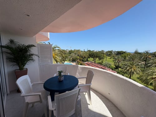 Balcón del apartamento SACORO en Marbella con vistas al mar, mesa de comedor, sillas blancas, palmeras y paisaje verde