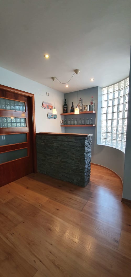 Barra de bar moderna con acabado en piedra oscura, estantes de madera, botellas e iluminación colgante en Villa Castelló Salou