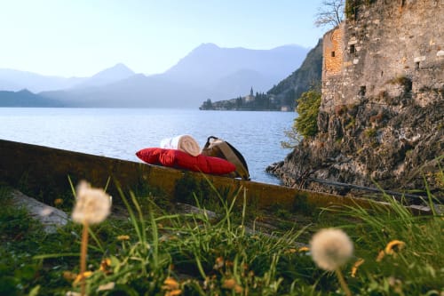 Persona che si rilassa su un cuscino rosso con vista sul Lago di Como a Varenna, con montagne ed edifici storici affacciati sul waterfront sullo sfondo