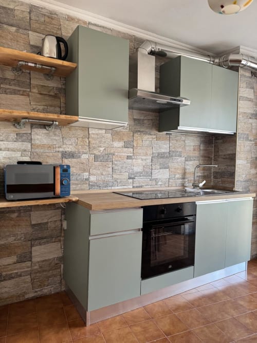Modern kitchen in Sormano apartment with sage green cabinets, stone backtile, wooden shelves, and stainless steel appliances