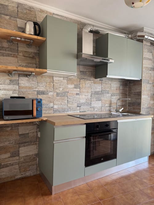 Modern kitchen in Sormano apartment with sage green cabinets, stone backtile, wooden shelves, and stainless steel appliances